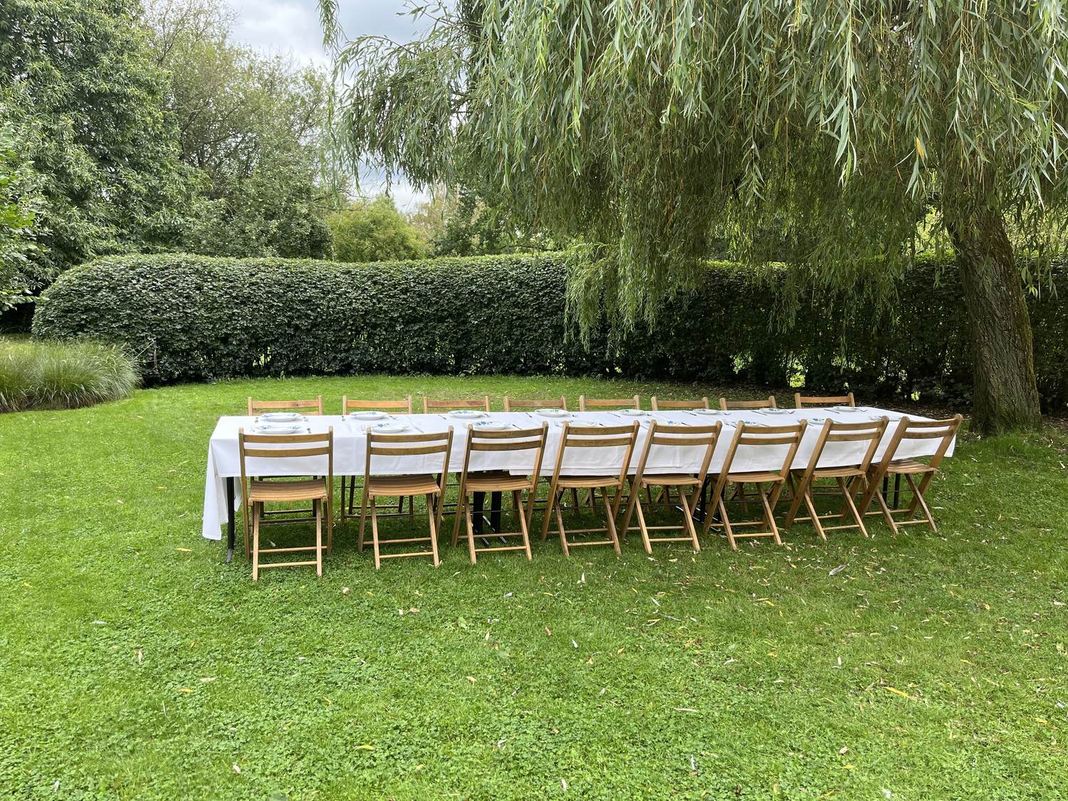 Lange gedekte tafel met houten klapstoelen onder een treurwilg in de tuin